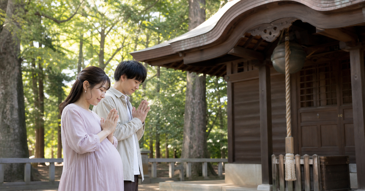 緑豊かな神社の社殿の前で、手を合わせて安産を祈る妊婦さんと夫